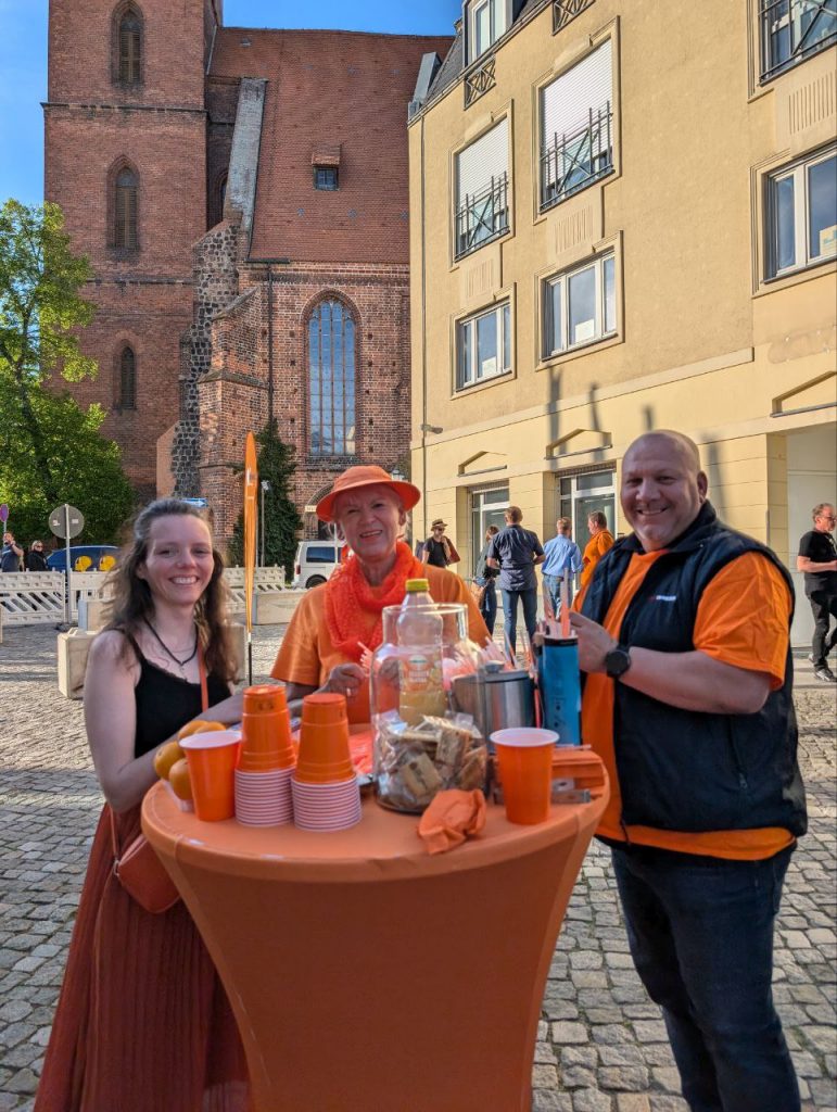 Nadine Schnittke, Christiane Strese und Alex Morzinek stehen an Tisch mit Orangensaft und Werbematerial