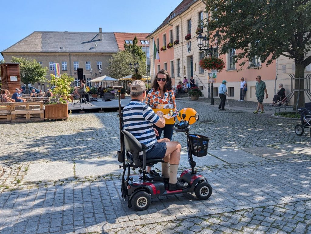 Nadine Schnittke verteilt Flyer und Orange an einen Rollstuhlfahrer auf Marktplatz